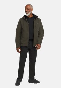 Olijfgroene fleece jas met een capuchon, gewoven stof en een ritssluiting aan de voorkant. Draagt over een zwart shirt, gecombineerd met zwarte broek en schoenen.