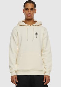 Cream hoodie with a front pocket, drawstring hood, and a cross design on the left chest. Made from soft fabric with ribbed cuffs.