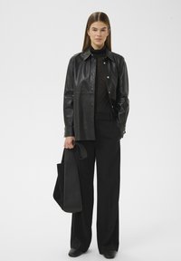 Schwarze Lederhemdjacke mit Druckknöpfen, getragen über einem schwarzen Rollkragenpullover und weiten schwarzen Hosen, kombiniert mit einer schwarzen Tragetasche.