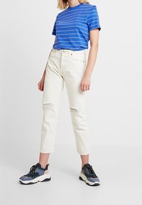 Niebieska koszulka w paski z krótkim rękawem połączona z kremowymi, podartymi dżinsami oraz wielokolorowymi masywnymi sneakersami z czarnymi sznurówkami i teksturowaną podeszwą.