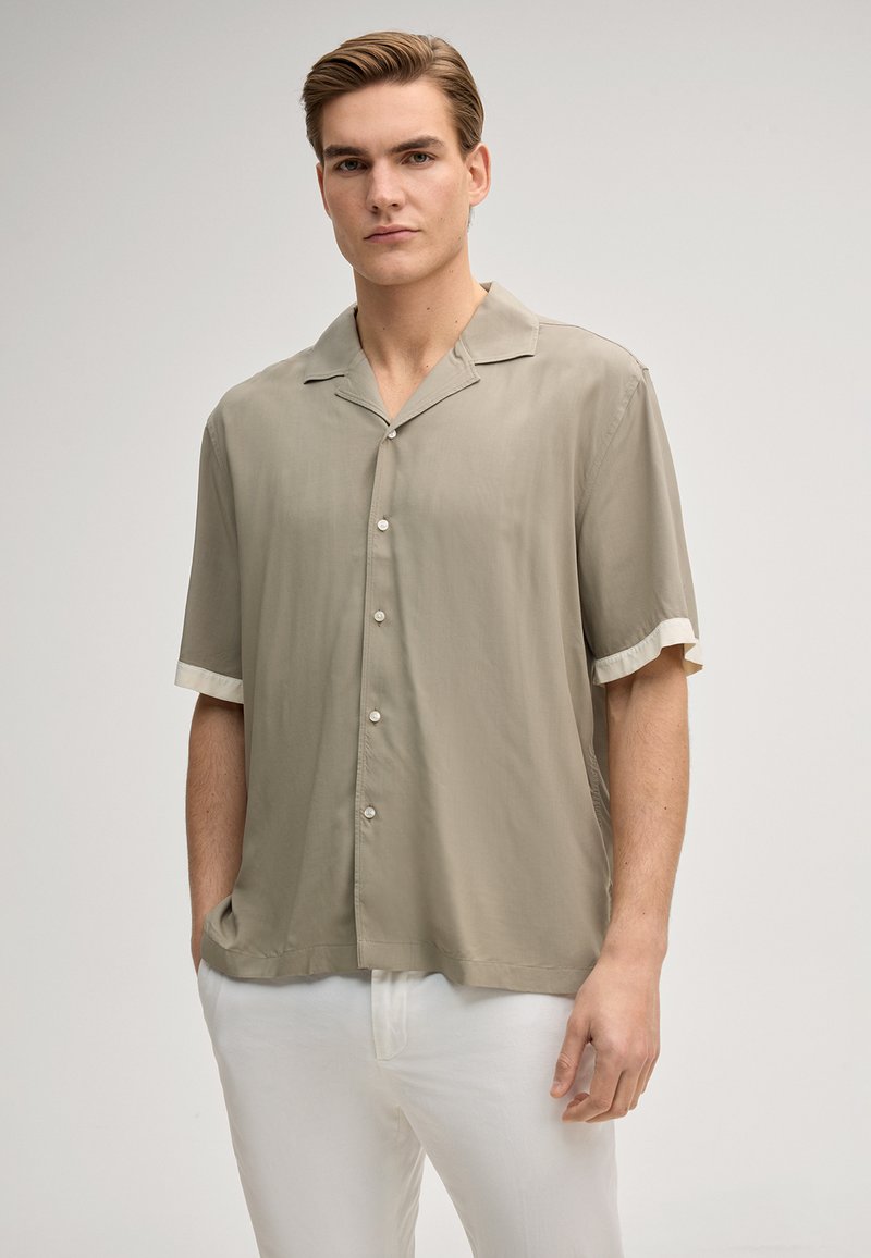Homme portant une chemise beige à manches courtes avec des bordures blanches aux manches et un pantalon blanc, debout devant un fond clair uni.