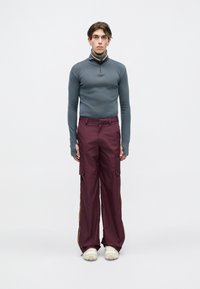 Mladenič v sivem dolgih rokavih z zadrgo in ohlapnih maroon cargo hlačah ter belih supergah, stoji ob enotni ozadju.