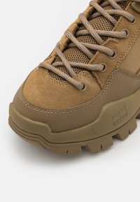 Brązowe zamszowe i siateczkowe trampki z teksturowaną gumową podeszwą, wzmocnionym czubkiem i płaskimi sznurówkami. Wyróżniające się detale to logo z boku.