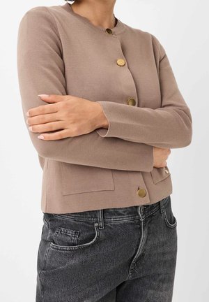 Vrouw in een beige cardigan met gouden knopen met gekruiste armen, gecombineerd met een zwarte, hooggetailleerde spijkerbroek tegen een effen achtergrond.