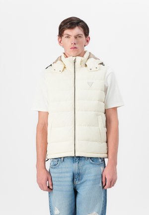 Padded cream vest with a high collar and zip closure, featuring shoulder snaps and two front pockets, worn over a white shirt and blue jeans.