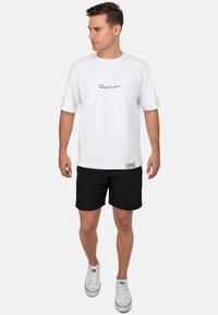 T-shirt bianco in cotone con un logo cursivo nero, abbinato a pantaloni neri e scarpe da ginnastica bianche. Design semplice con maniche corte e linee pulite.