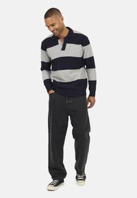 Pull en tricot rayé gris et marine, avec un col et une coupe décontractée, associé à un pantalon baggy noir et des baskets noires.