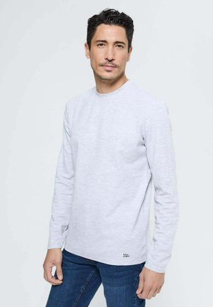 T-shirt à manches longues gris clair en mélange de coton doux ; présente un col rond et un petit logo en bas à gauche. Porté avec un jean bleu.