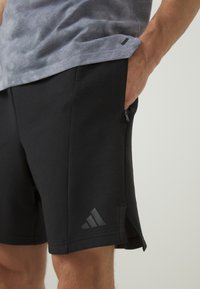 Shorts de sport noirs en tissu flexible ; dotés de poches latérales et d'un petit logo gris au bord. Longueur courte avec des fentes sur les côtés.