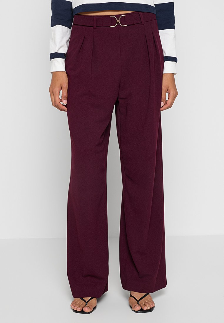 Wijdvallende broek in donkere bordeauxstof, met voorplooien en een gouden riemgesp accent. Gladde textuur en tailored fit.