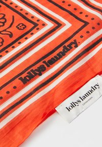 Oransje skjerf med kraftige svarte mønstre, stripete kanter og et merke som sier "lolly's laundry" i svart på hvit bakgrunn.
