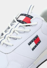 Biele kožené tenisky s bielymi šnúrkami, ťahacím pútkom s modrými pruhmi a logom Tommy Jeans v červenej, bielej a námorníckej modrej farbe.