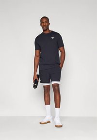 Άνδρας σε μαύρη αθλητική ενδυμασία κρατώντας μαύρη φιάλη νερού, φορώντας λευκές κάλτσες και λευκά αθλητικά παπούτσια, στέκεται μπροστά σε απλό γκρι φόντο.