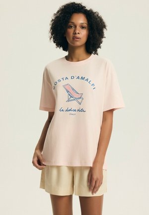 Kobieta z kręconymi włosami w jasnoróżowej koszulce z grafiką pasiastych leżaków plażowych i napisem "Costa D'Amalfi la dolce vita Włochy".