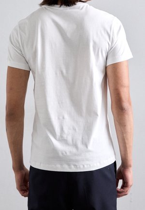 Homme portant un t-shirt blanc uni à manches courtes et un pantalon noir, vu de dos sur un fond clair uni.