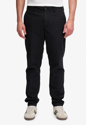 Man draagt zwarte casual broek en witte sneakers, staat tegen een effen lichte achtergrond, handen ontspannen langs het lichaam.