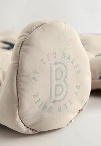 Beige gepolstertes Kissen mit geprägtem Logo "B" und dem Text "TED BAKER" in Hellblau. Glatte Oberfläche mit abgerundeten Kanten.