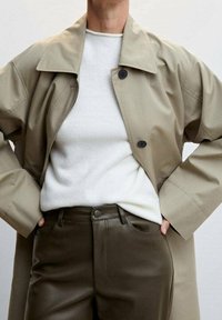 Beiger Trenchcoat mit großem Kragen und schwarzen Knöpfen, getragen über einem weißen Pullover und dunkelgrünen, glatt verarbeiteten Hosen.