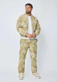 Homme debout portant une veste à motifs floraux verts et beiges et un pantalon assorti, avec des baskets blanches et un t-shirt blanc.