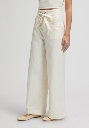 Kobieta ubrana w kremowe spodnie o wysokim stanie i szerokich nogawkach z przednimi kieszeniami oraz paskiem do wiązania, w połączeniu z beżowymi płaskimi butami i krótkim topem.