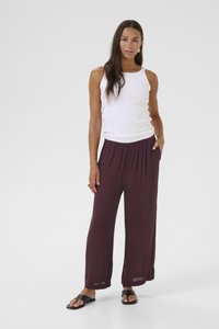 Woman standing with one hand in pocket wearing a white sleeveless top, loose maroon pants, and black slide sandals on a white background.