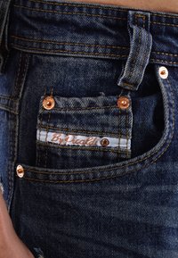 Dunkelblaue Denim-Jeans haben kupferne Nieten, orangefarbene Nähte und eine kleine Fronttasche mit einem weißen Label, auf dem "By Piccoli" steht.