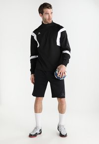 Sweatshirt de sport noir avec des accents blancs, col zippé à moitié, associé à un short noir. Le modèle tient un ballon de handball, portant des chaussures de football et des chaussettes blanches.