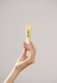 Gul tube med Torriden læbesome heldt af en hånd. Har en tekstureret overflade og en hvid bund med klare sorte bogstaver.