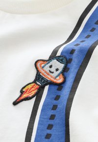 White cotton fabric featuring a blue striped design with black lines and dotted accents. Embroidered rocket design in colors orange, blue, and yellow.