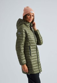 Olijfgroene pufferjack met capuchon, ritszakken, een gequiltte textuur en een ronde logopatch op de mouw. Draagt een roze beanie.