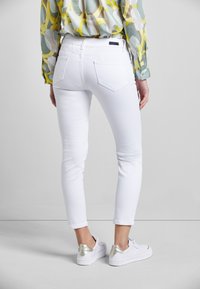 Frau in weißen, engen Jeans, weißen Sneakers mit silbernen Absätzen und einer gemusterten Bluse mit langen Ärmeln steht vor einem einfarbigen Hintergrund.