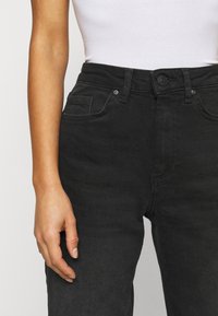 Mustat denimpantterit, joissa on korkea vyötärö, napitus ja etutaskut. Kankaan pinta on sileä ja siinä on hienovaraisia ompelut yksityiskohtia.