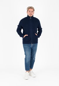 Maglione navy con cerniera e collo alto, texture a coste e tasche frontali. Abbinato a jeans azzurri a gamba stretta e sneakers bianche.