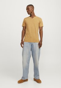 Camiseta de corte corto en color mostaza con escote en V y detalle de dos botones, combinada con jeans de un azul claro y zapatos marrones tipo mocasín.
