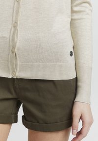 Beige cardigan laget av myk strikk, med en rad runde knapper og ribbestrikkede mansjetter. Kombinert med rullede olivengrønne shorts.