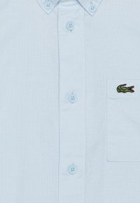 Camicia button-up celeste realizzata in tessuto con finitura testurizzata. Presenta una tasca applicata con un logo di coccodrillo verde e bottoni abbinati.