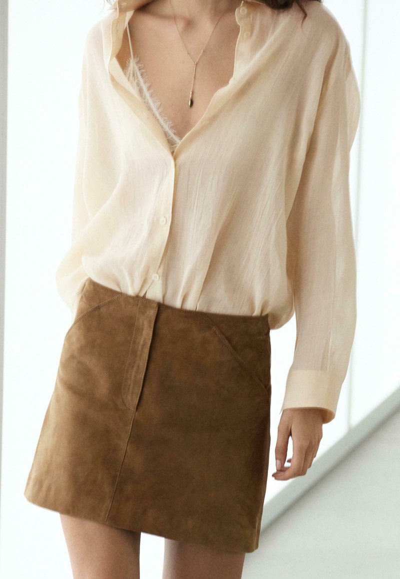 Kvinde iført en gennemsigtig cremefarvet skjorte med knapper over en hvid blonde-camisole, kombineret med en brun ruskind mini nederdel.