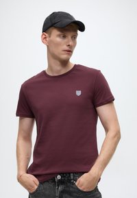 Tumepunane lühikese varrukaga T-särk ümarkaelusega, millel on väike tikitud kilbilogo. Sobitatud tumedate teksapükstega ja musta nokamütsiga.