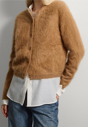 Frau trägt eine flauschige braune, geknöpfte Strickjacke über einem weißen Hemd und blauen Jeans, steht mit einer Hand in der Tasche vor einer schlichten Wand.