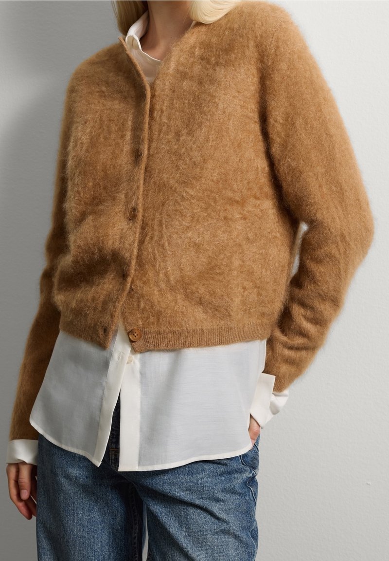 Femme portant un cardigan marron pelucheux boutonné sur une chemise blanche et un jean bleu, debout avec une main dans la poche contre un mur uni.