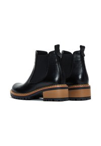 Botas de tobillo de cuero negro con paneles elásticos laterales, punteras redondeadas y un tacón grueso de color marrón claro. Cuentan con una suela texturizada con patrones de agarre.