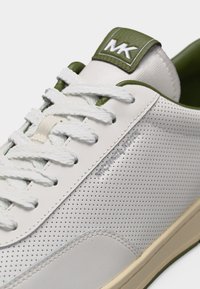 Κοντινό πλάνο σε λευκό πάνινο παπούτσι με τρύπες, λευκά κορδόνια, γιακά ελιάς και λογότυπο «MK» στη γλώσσα.