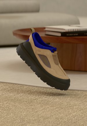 Chaussure UGG beige et bleue à enfiler, avec semelle épaisse noire, flottant au-dessus d'un tapis tissé, d'une table basse en bois et d'un canapé beige en arrière-plan.