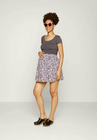 Camisa ajustada de manga curta cinza, saia curta de padrão floral em roxo claro e sabrinas de madeira pretas. Modelo usando óculos de sol, em pé com uma mão no quadril.