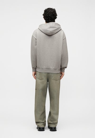 Άνδρας με σγουρά μαλλιά, φοράει γκρι φούτερ με κουκούλα και φαρδιά ξεθωριασμένα πράσινα παντελόνια, στέκεται με την πλάτη προς το μέρος μας σε απλό φόντο.
