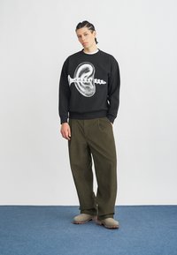 Črna jopa z velikim belim motivom vijaka in školjke. Stilizirana z ohlapnimi hlačami olivno zelene barve in bež obuvali.