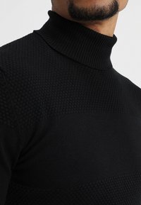 Zwarte coltrui met een textuur gebreid patroon, voorzien van een geribde kraag en afwisselende patronen, ontworpen voor warmte en comfort.