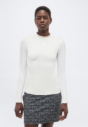 Žena oblečená v bielom športovom tričku s dlhými rukávmi a krátkej čiernej sukni s bielo-modrými geometrickými vzormi, stojaca pred jednoduchým pozadím.