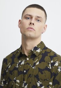 Camisa de hombre en estampado floral verde y marino, con cierre de botones en la parte delantera, cuello doblado y un bolsillo en el pecho; tejido de algodón con textura suave.