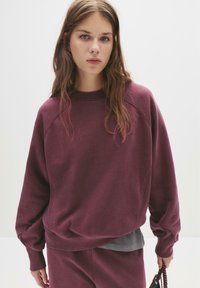 Felpa oversize bordeaux con collo a girocollo a costine, maniche raglan e polsini elasticizzati. Tessuto morbido, indossata sopra una maglietta grigia.